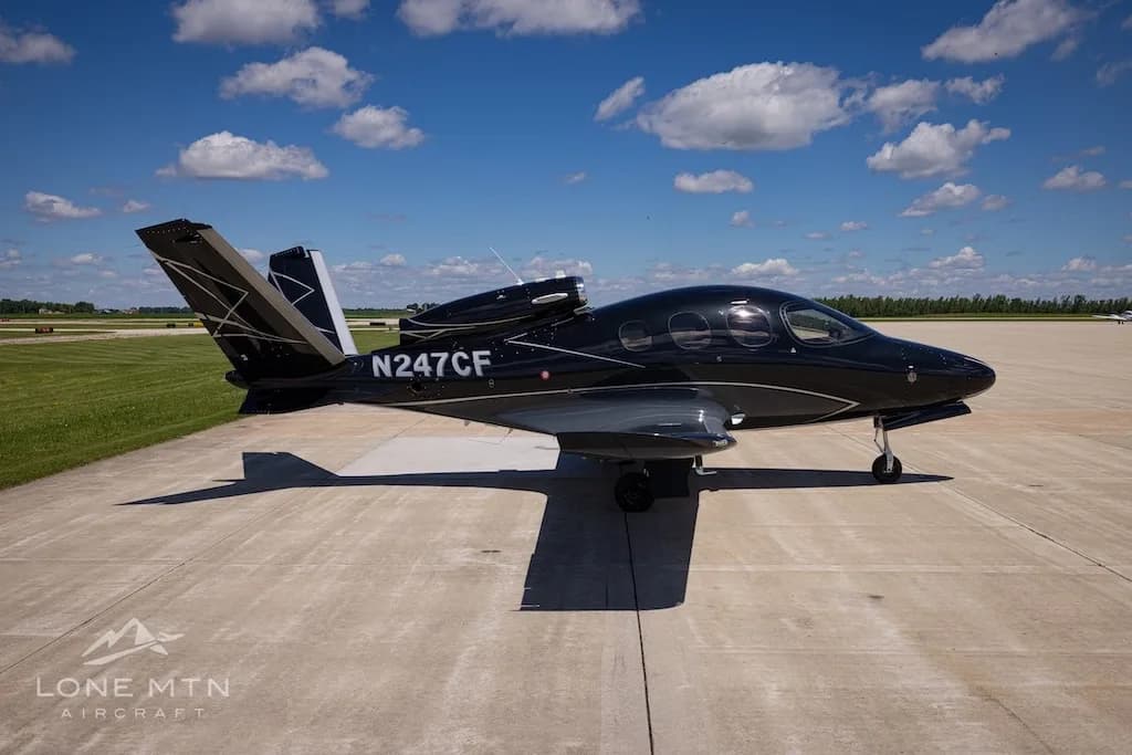 A black Cirrus Vision Jet (N247CF) with silver lower fuselage and wings parked on a concrete surface under a partly cloudy sky.