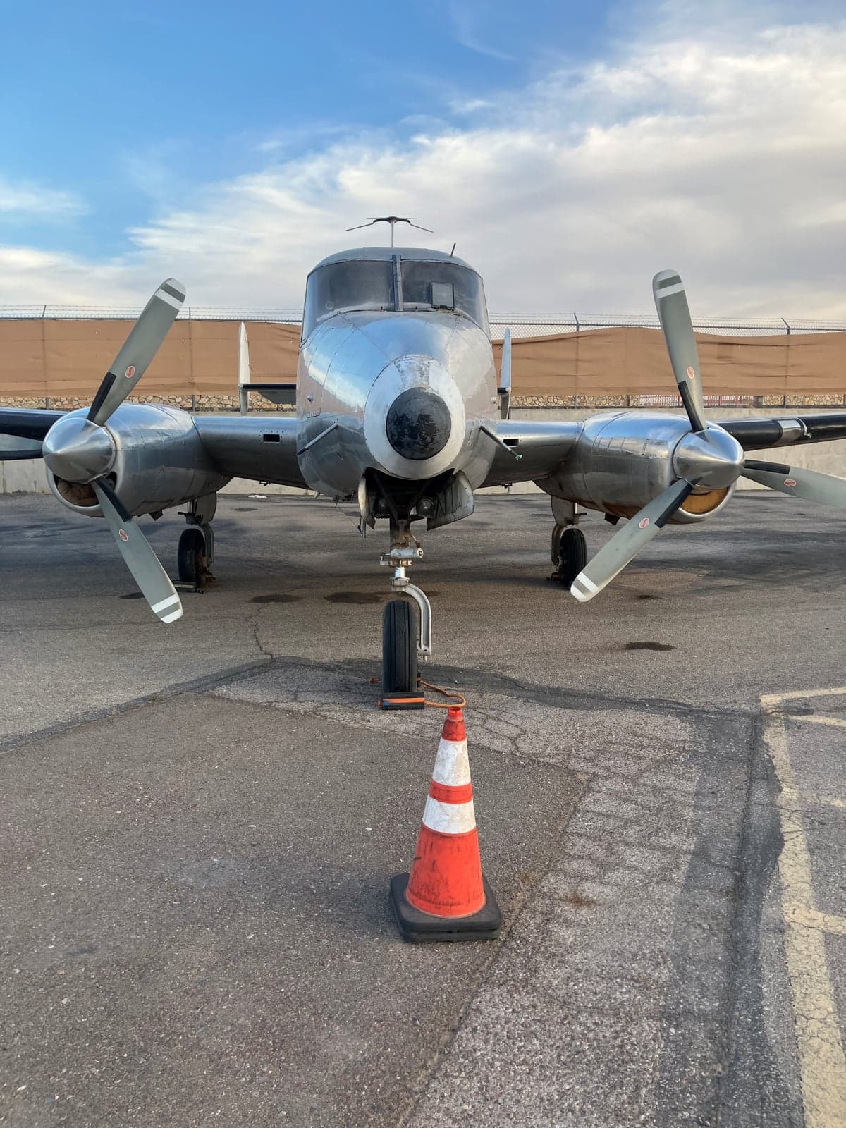 Front view of a silver Twin Bonanza aircraft on the tarmac with a safety cone in front of the nose gear.