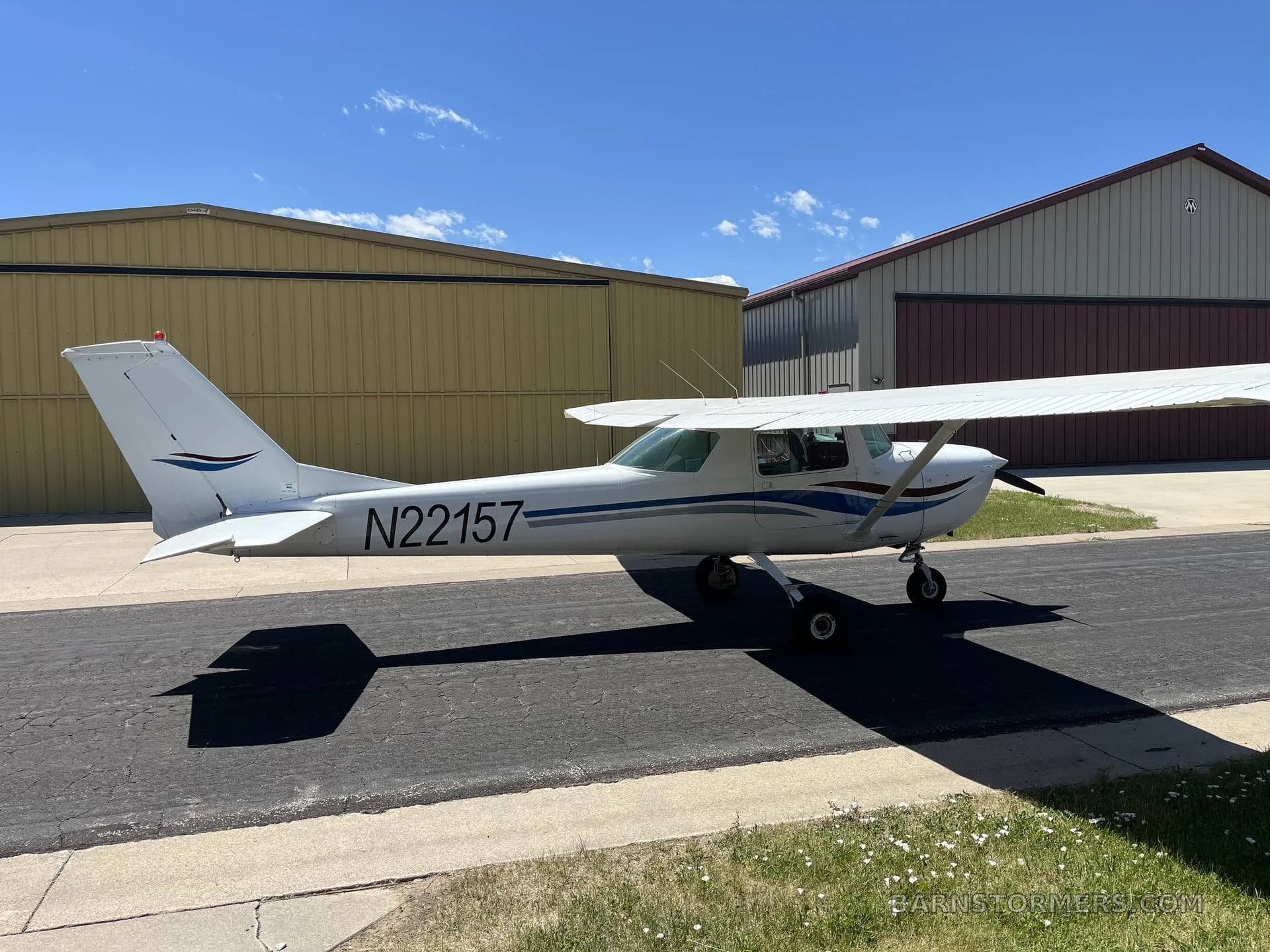 A white Cessna 150/152 type aircraft with registration N22157 and blue and red stripes parked on an asphalt ramp between hangars.