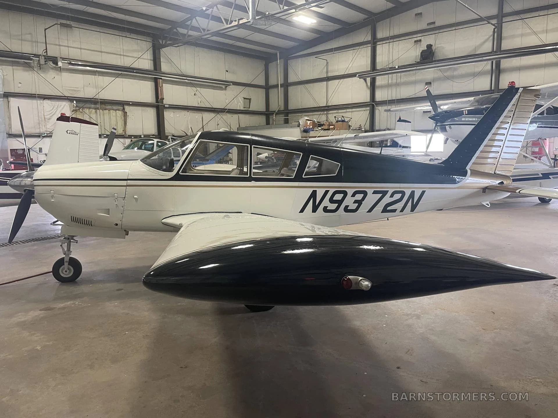 A white and black single-engine aircraft with a gold stripe, registration N9372N, parked in a hangar, viewed from the front-left.