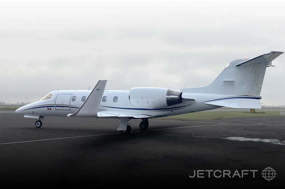 A white business jet with a black and gold stripe and a Mexican flag on the nose, parked on a runway under an overcast sky.