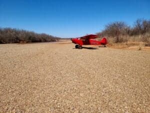 A red aircraft is parked on a gravelly surface near some trees.