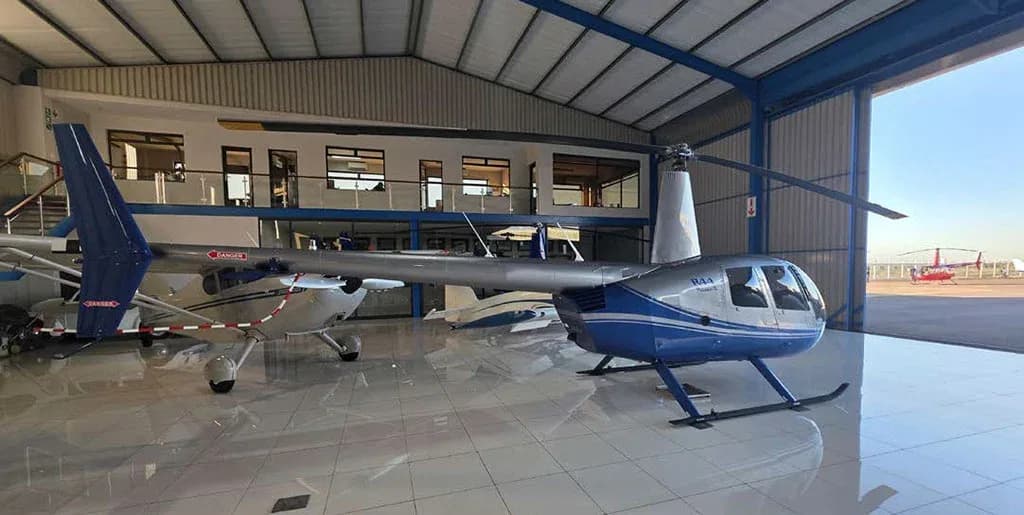 A silver and blue Robinson R44 Raven II helicopter in a hangar with the door open to the tarmac and another red helicopter outside.