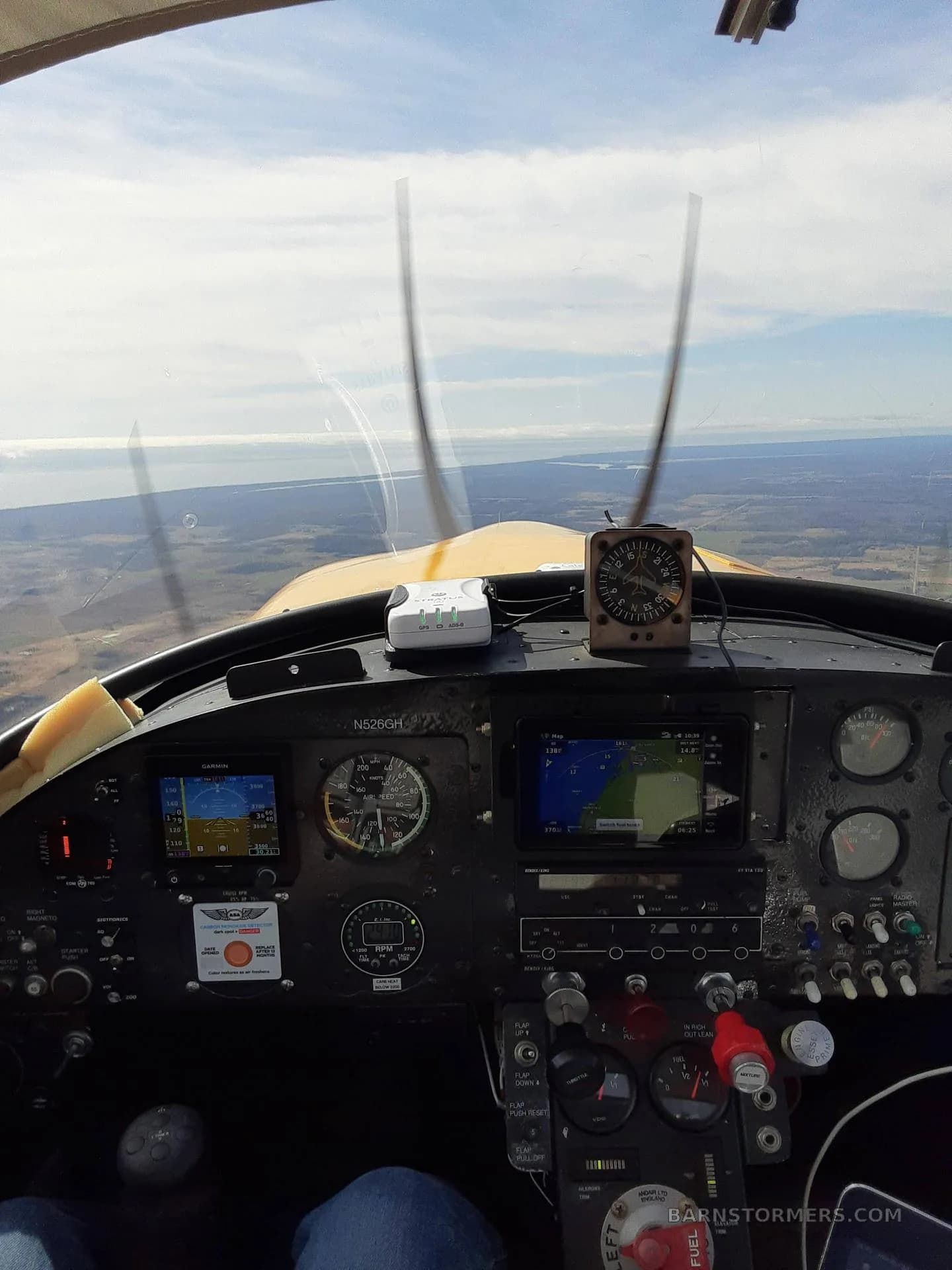 View from the cockpit of a yellow aircraft in flight, showing the instrument panel with a Garmin display and the landscape below.