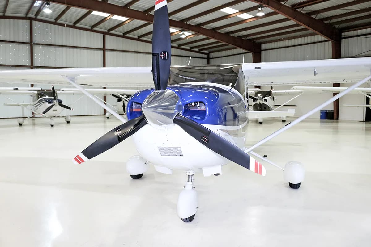 A head-on view of a white and blue single-engine propeller aircraft with a three-bladed propeller, parked in a hangar.