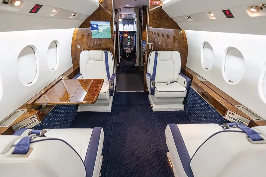A wide view of a private jet cabin with white and blue seats, a wooden table, and a screen displaying a map.