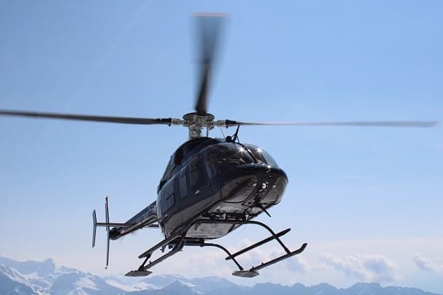 A black Bell 407GX helicopter in flight against a blue sky and snowy mountains.