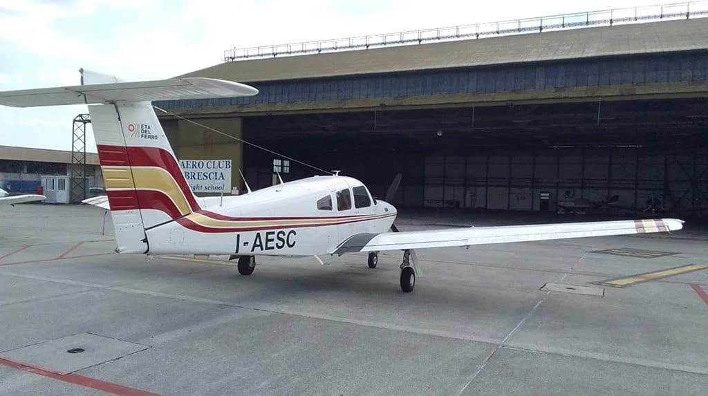A white Piper PA-28 aircraft with red and gold stripes parked on an airfield in front of a large hangar.