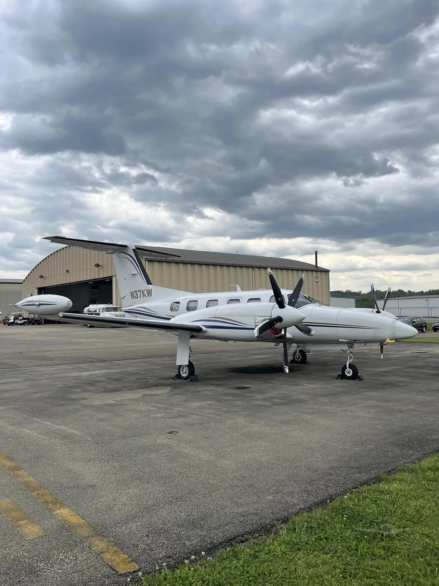 Three-quarter front view of a white twin-engine turboprop aircraft (N37KW) with blue and gold stripes on the tail, parked near a hangar.