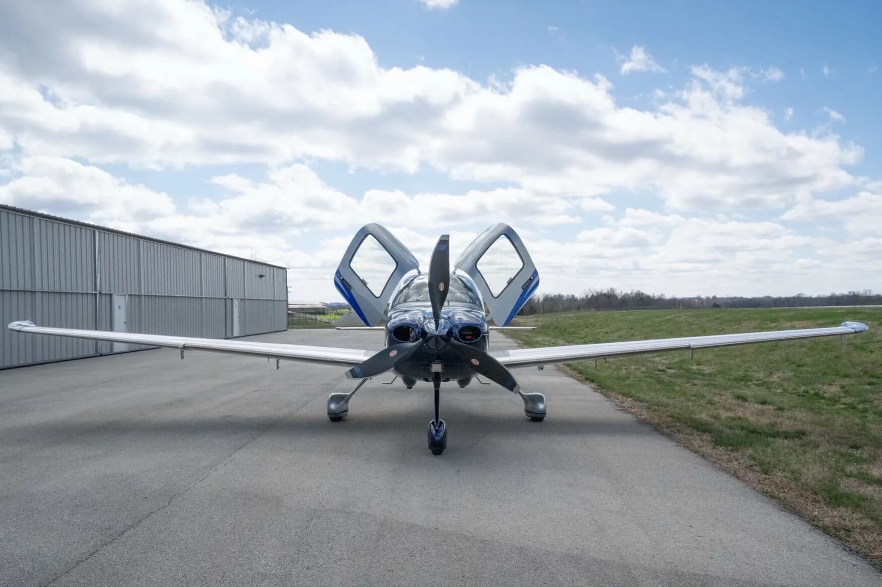 Front view of a silver and blue Cirrus SR22T aircraft with its gull-wing doors open, parked on an airfield.