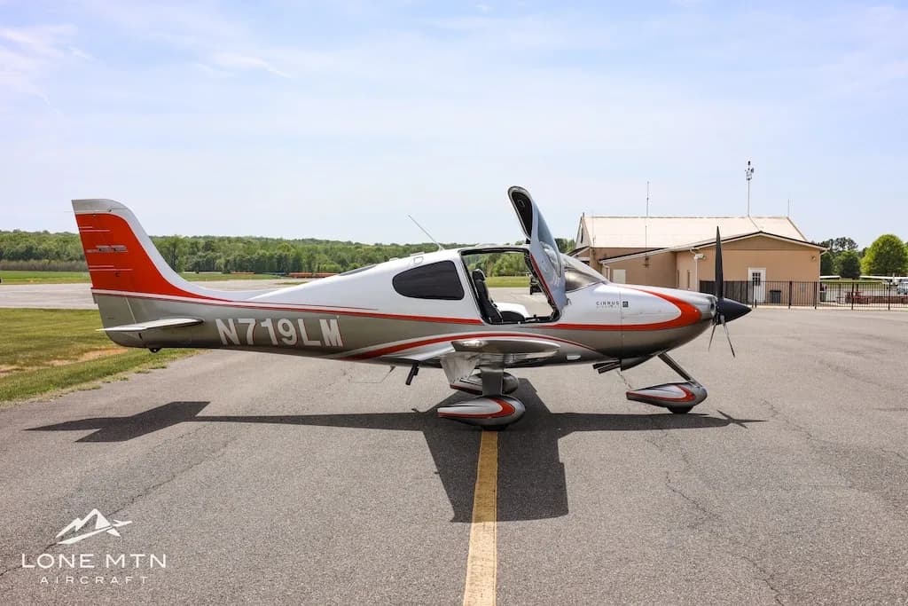 Side view of a silver and orange Cirrus SR22 aircraft with the pilot's door open, parked on an asphalt runway.