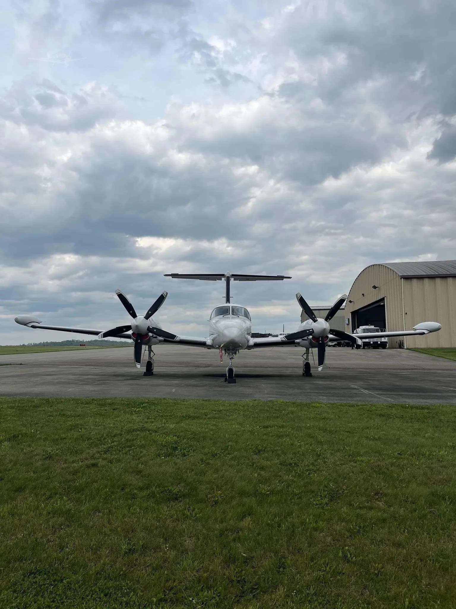 Front view of a white twin-engine turboprop aircraft with black propellers, parked in front of a hangar under a cloudy sky.