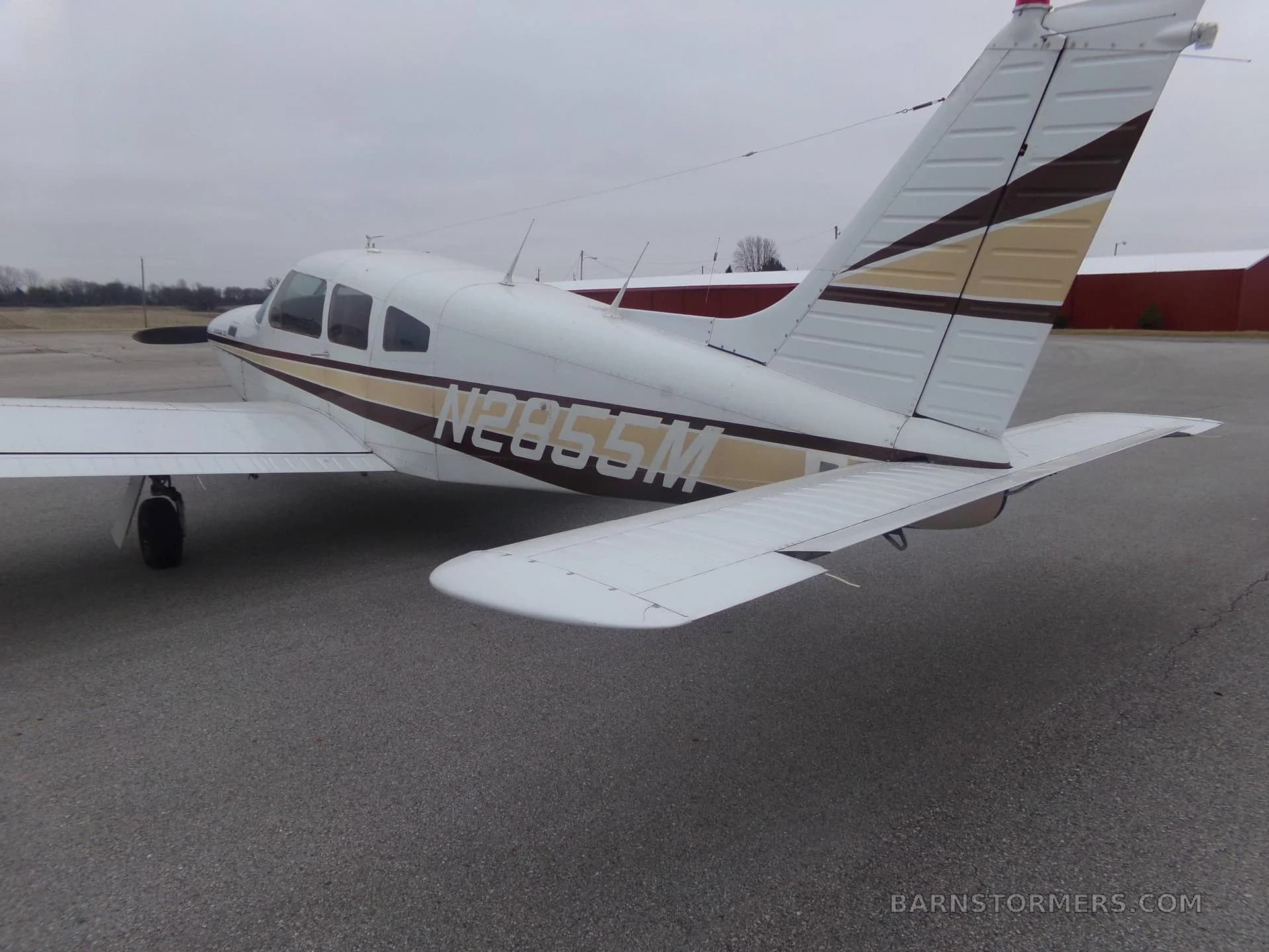 The rear-right view of a white Piper Arrow III aircraft with brown and gold stripes, registration N2855M, parked on an asphalt tarmac.
