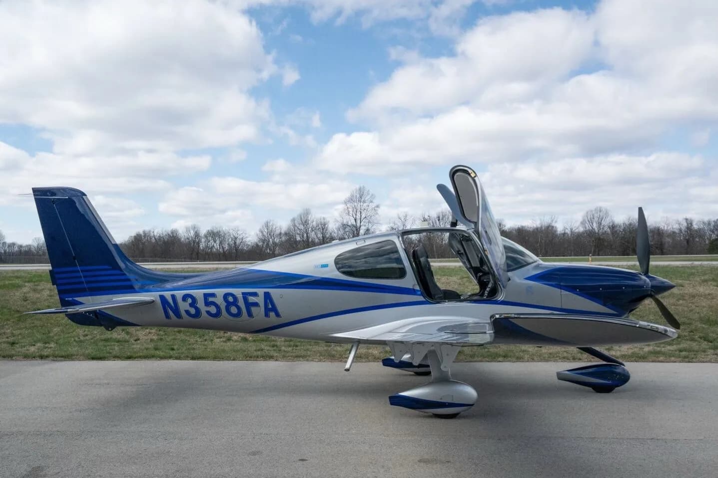 A silver and blue Cirrus SR22T aircraft, registration N358FA, with its gull-wing doors open, parked on an airfield.