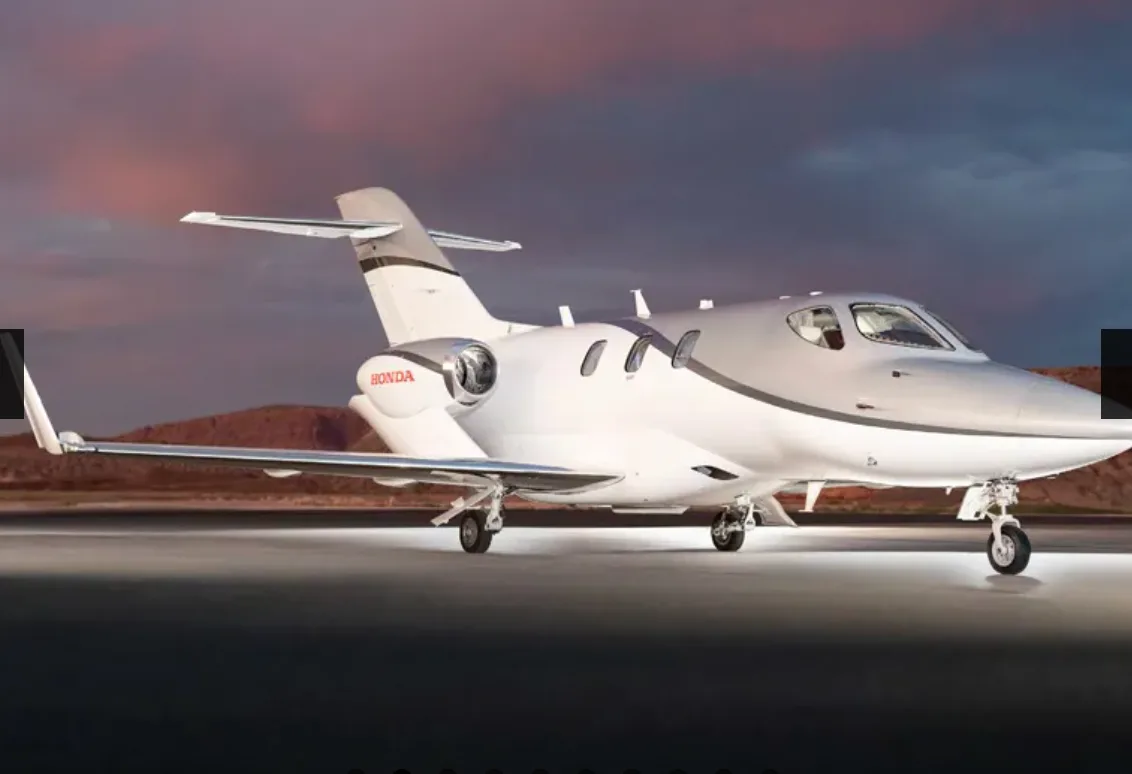 A HondaJet aircraft in a grey and white paint scheme, parked on a tarmac with mountains in the background at dusk.
