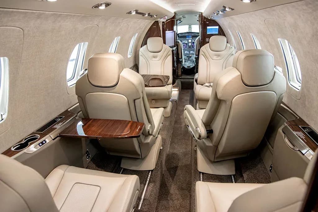The luxurious interior cabin of a private jet features light tan leather seats with pull-out tables, looking towards the cockpit.