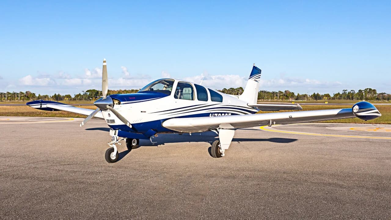 A white and blue Beechcraft Bonanza V-tail aircraft sits on an asphalt tarmac under a clear blue sky.