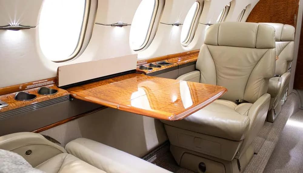 A close-up of a beige leather seat and a polished wooden table extended from the sidewall next to a window in a private jet.