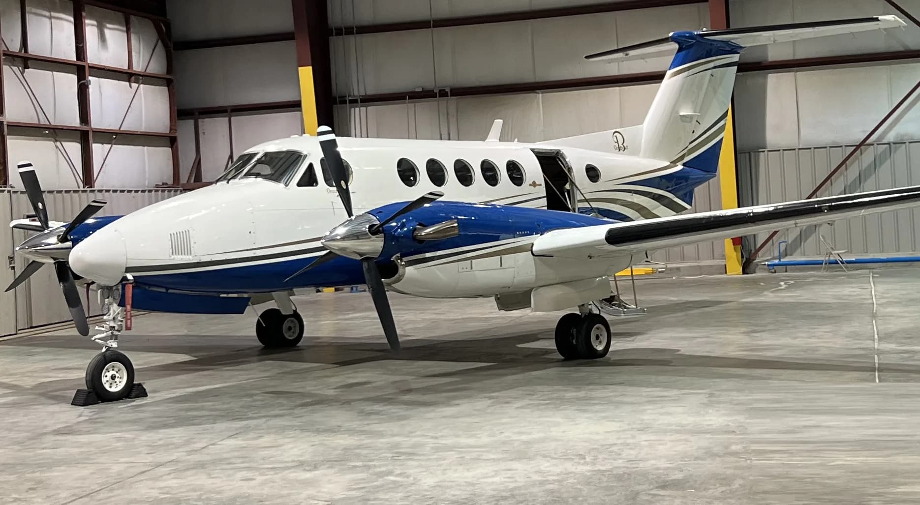 A white, blue, and gold twin-turboprop aircraft, possibly a King Air, is parked in a hangar with its main door open.
