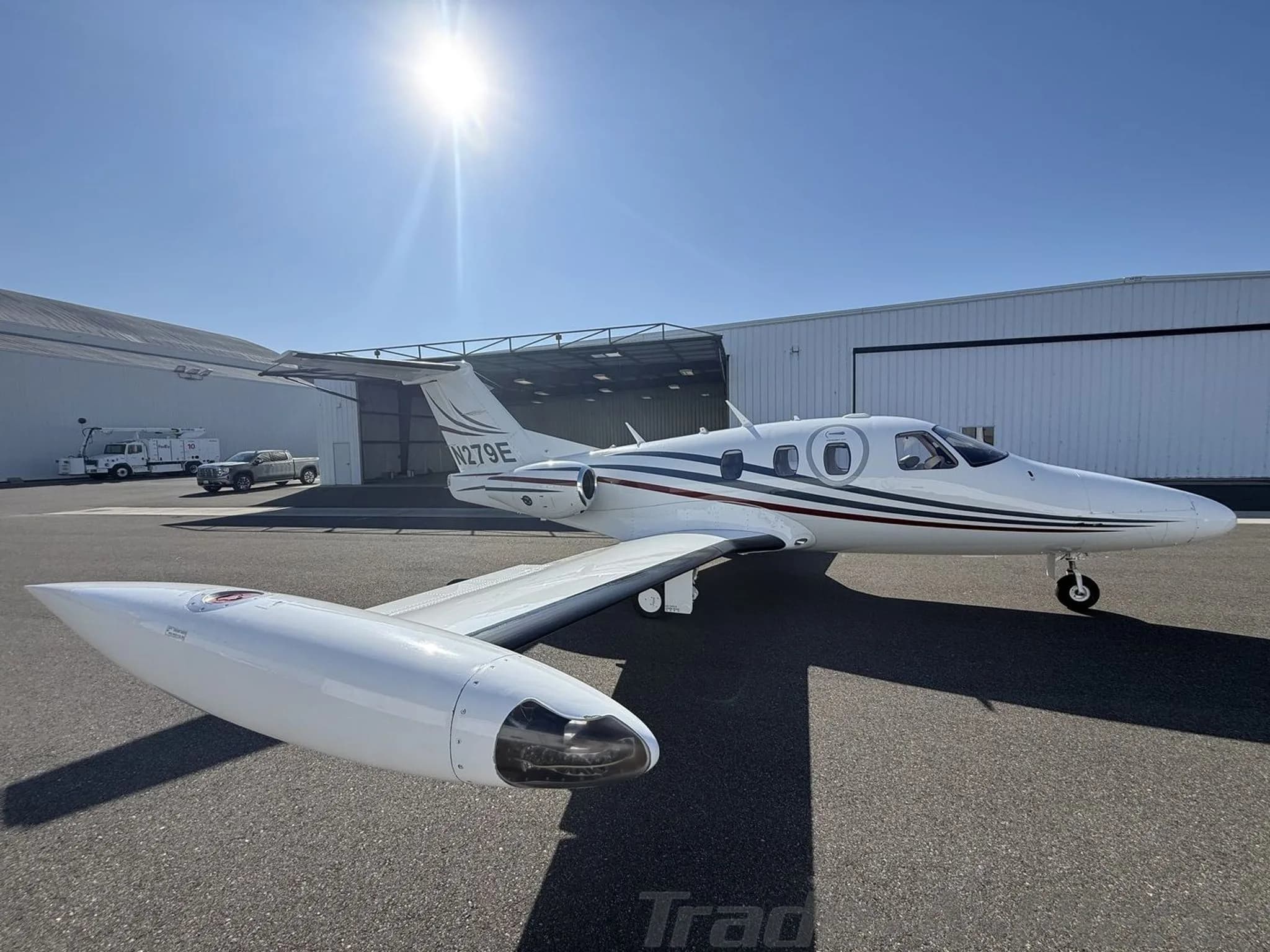 A white private jet with red and blue stripes and registration N279E parked on an apron under a sunny sky.
