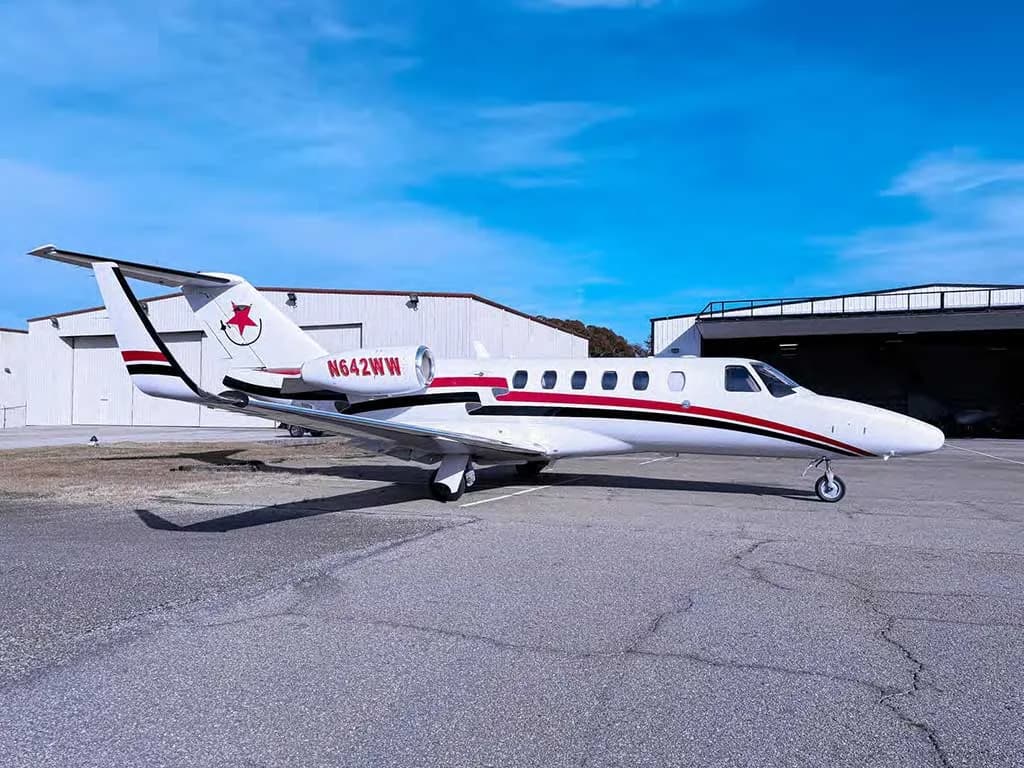 A white Cessna Citation private jet with red and black stripes and tail logo, parked on an asphalt tarmac in front of hangars.