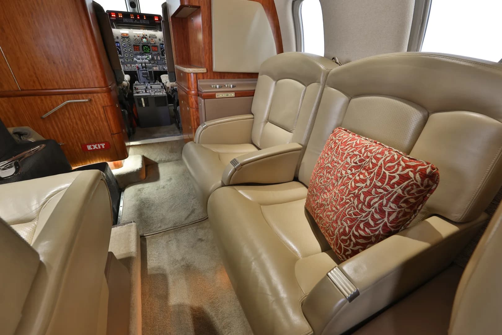A close-up view of the beige leather seats in a business jet cabin, with a red patterned pillow and a glimpse of the cockpit.