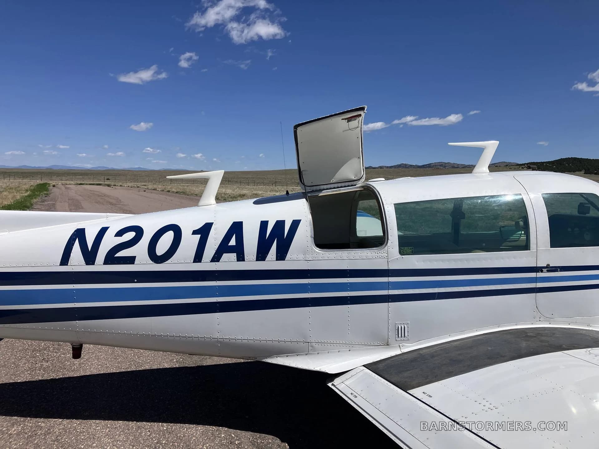 A close-up side view of a white Mooney M20J aircraft, registration N201AW, with its baggage door open.