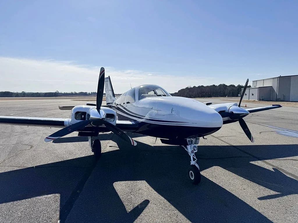 A white and blue twin-engine aircraft, likely a Beechcraft Baron, parked on an airfield, viewed from the front.