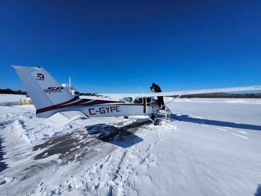 A white and red Cessna 172 (C-GYPE) is parked in a snowy field with a person inspecting its wing.