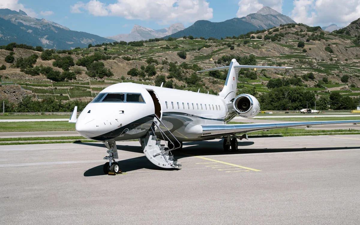 A white and dark grey business jet with its door open and stairs extended, parked on an airfield with mountains and vineyards in the background.