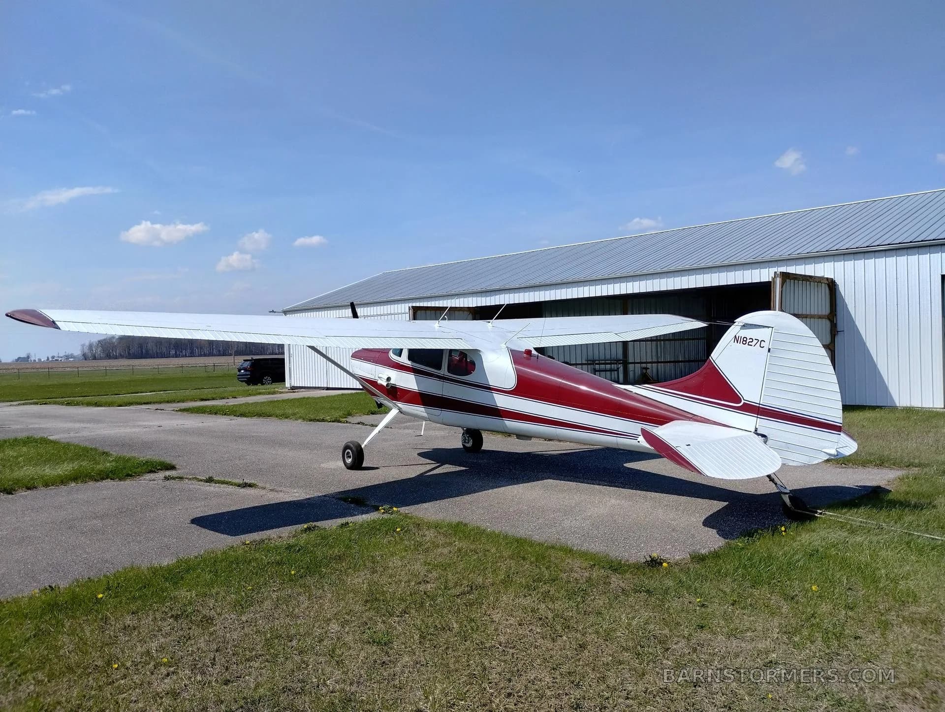 A red and white single-engine propeller aircraft with tail number N1827C is parked on an asphalt pad in front of an open hangar door.