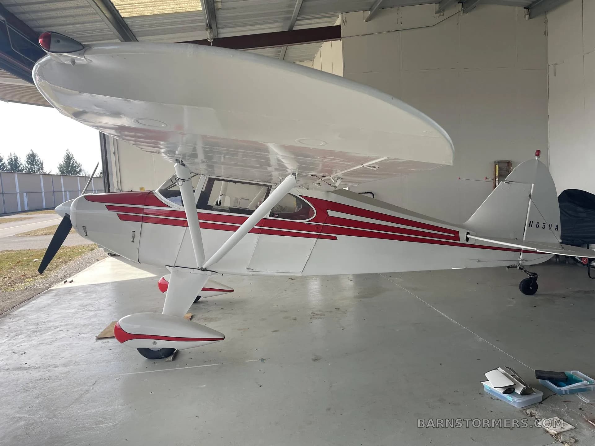 A white and red Piper PA-12 Super Cruiser aircraft is parked inside a hangar with the door open.