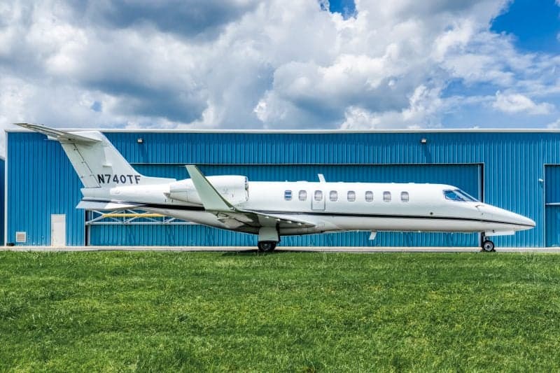 A white Learjet 75 with registration N740TF parked on green grass in front of a blue hangar under a cloudy sky.