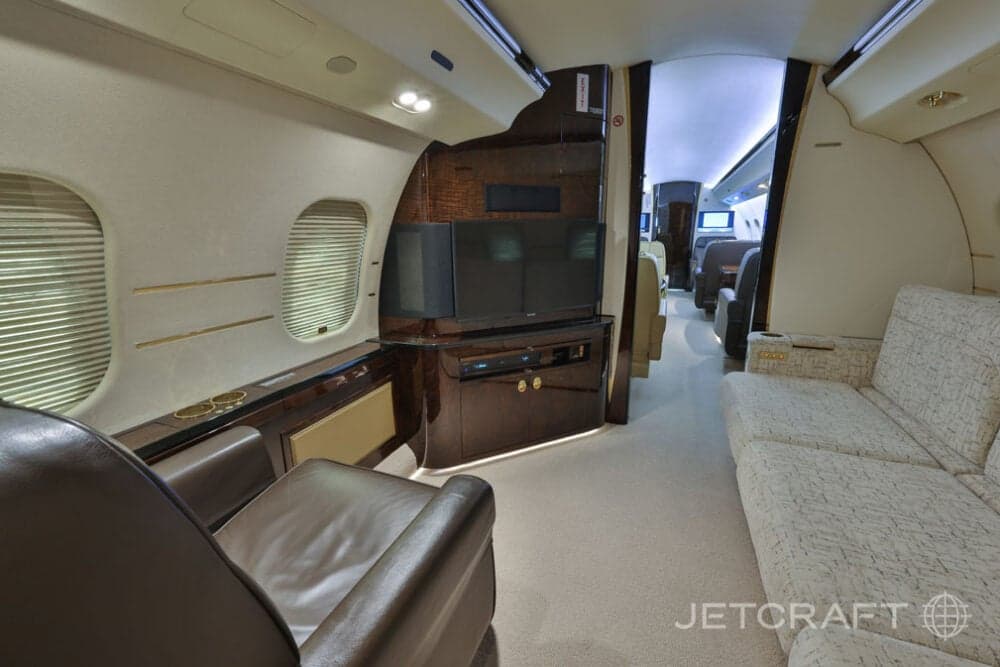 A section of the business jet cabin featuring a light-colored sofa and a dark wood entertainment console with two screens.