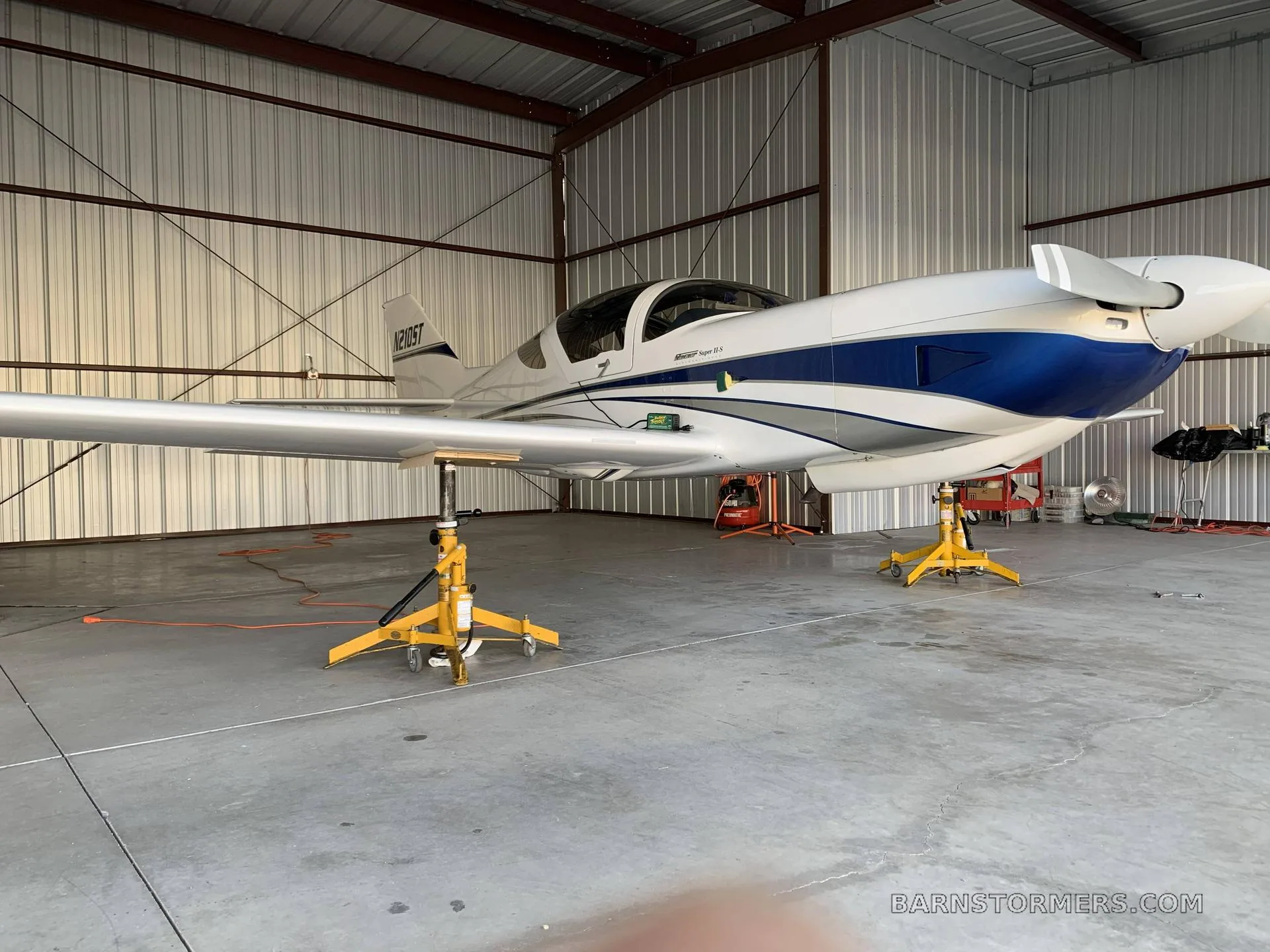 A white and blue single-engine aircraft (N210ST) is shown on maintenance jacks inside a hangar.