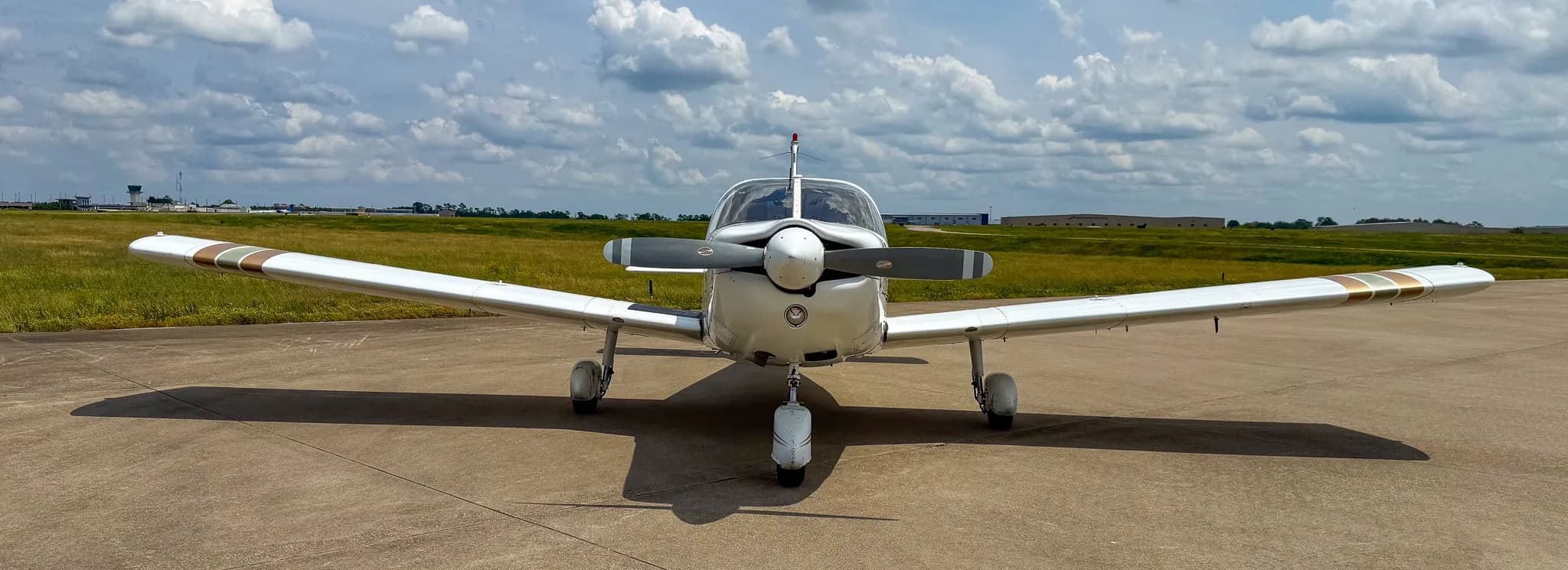 A front view of a white Piper Cherokee Six aircraft with brown and green stripes parked on an airfield.
