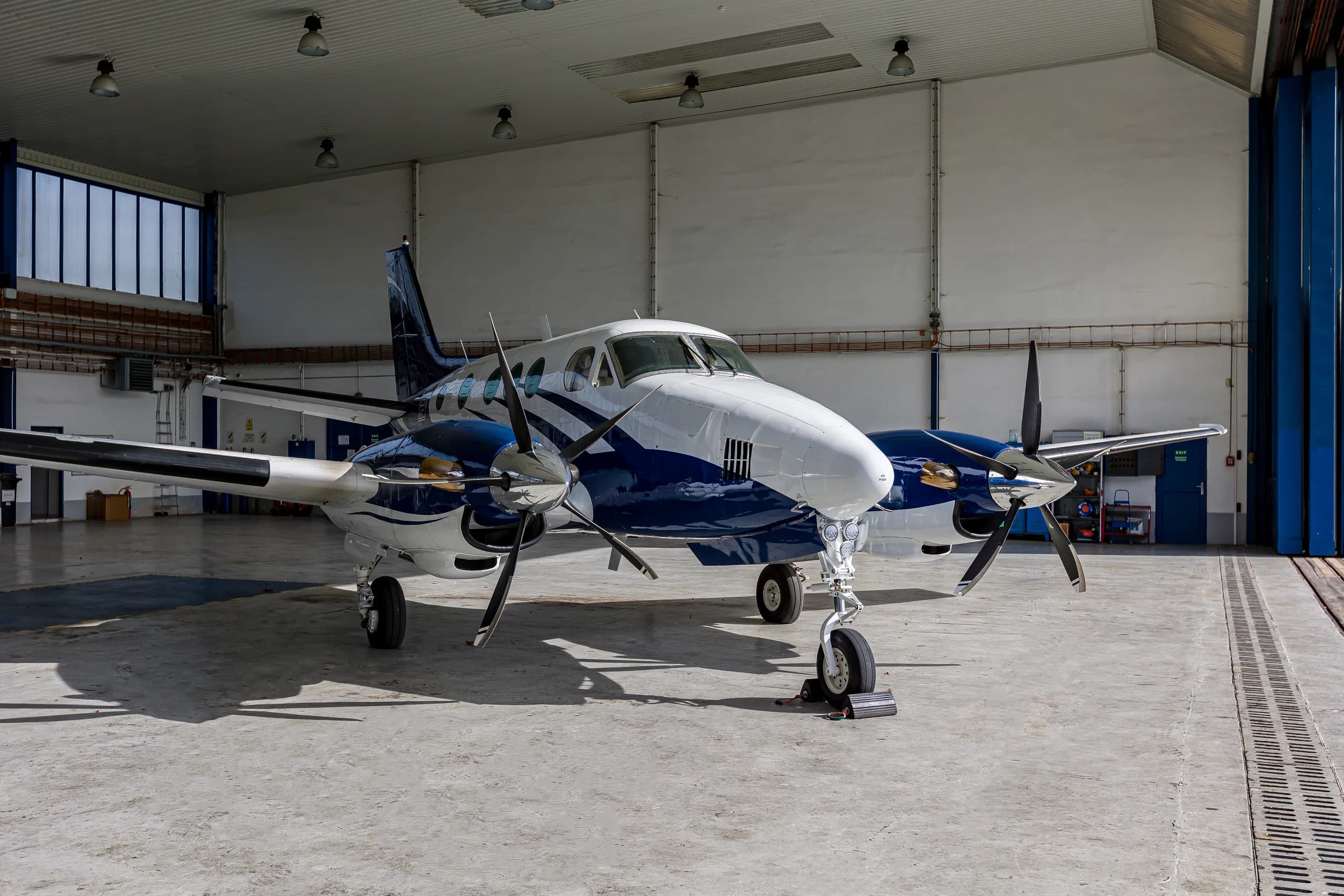 A front view of a white and blue twin-engine turboprop aircraft, likely a King Air, parked inside a hangar.