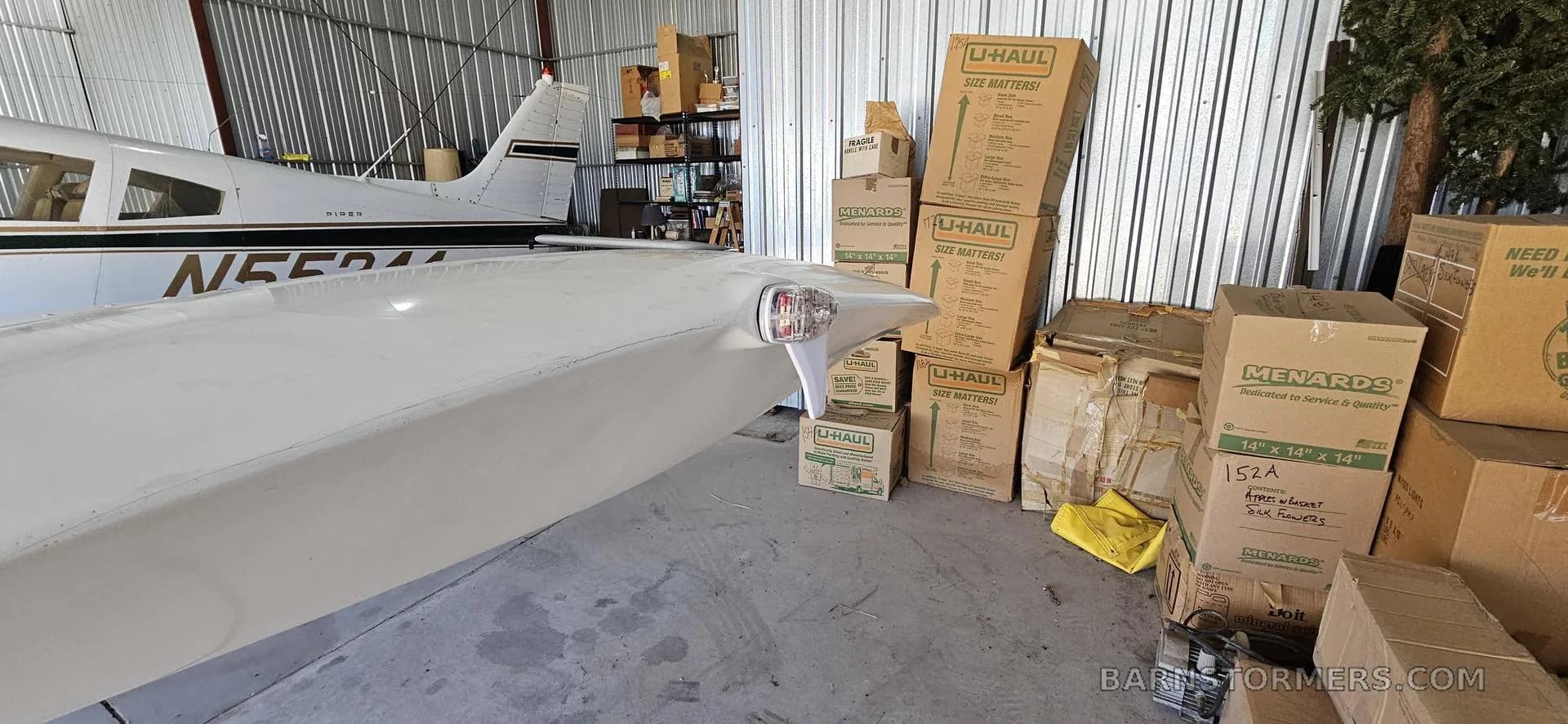 The white wingtip and part of the fuselage of a Piper aircraft (N55244) inside a hangar filled with boxes.