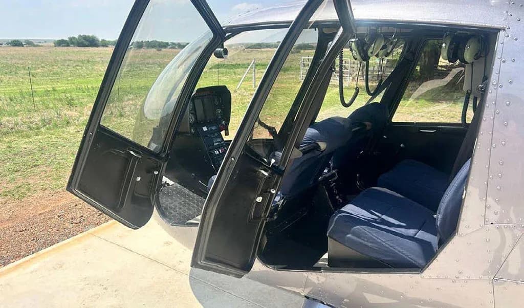 The open doors of a silver Robinson R44 helicopter revealing blue seats and a partial view of the cockpit.