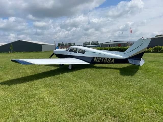 Side view of the N218SA single-engine aircraft, painted white and blue, parked on a grassy airfield.