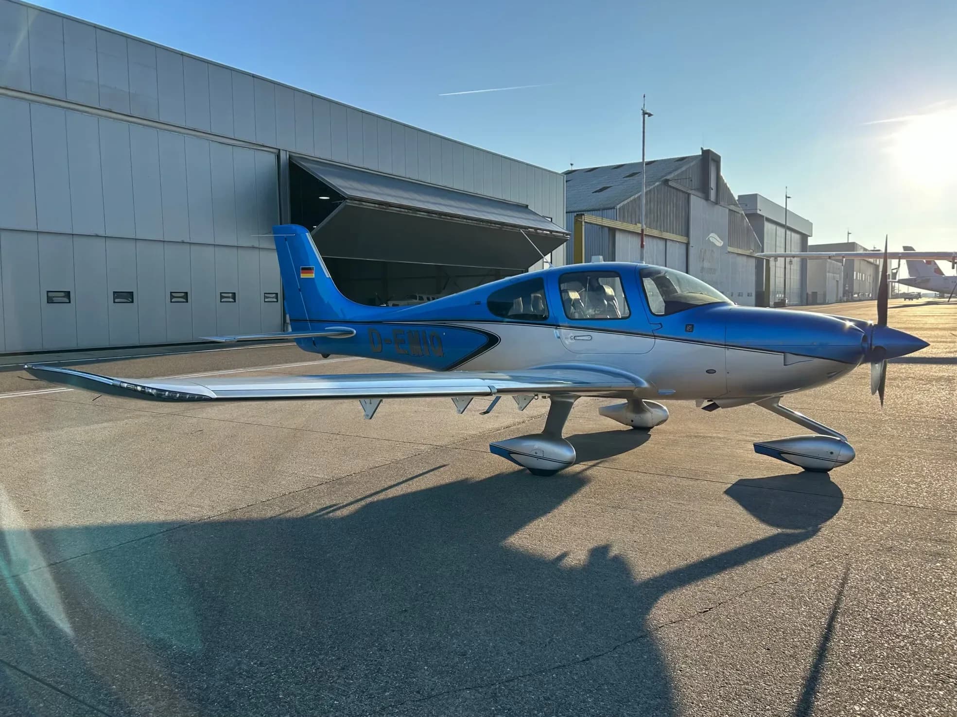 A blue and silver Cirrus SR22 aircraft parked on an asphalt apron with hangars in the background, bathed in late afternoon sun.