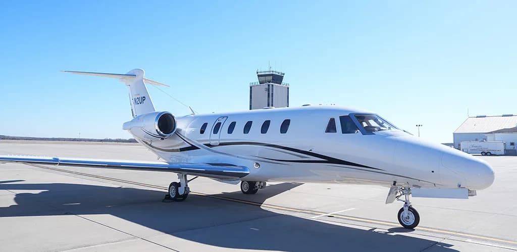A white Cessna Citation business jet with black and grey stripes, registration N2UP, parked on an airfield with a control tower in the background.
