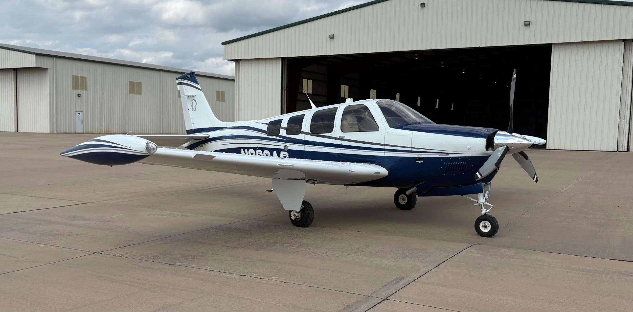 A white and blue Beechcraft Bonanza N686AG is parked on an airfield tarmac with a hangar in the background.