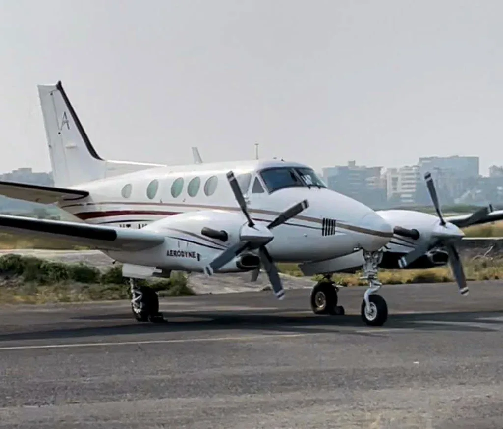 A white Beechcraft King Air C90B with brown and black stripes, parked on an airfield with a city skyline in the background.