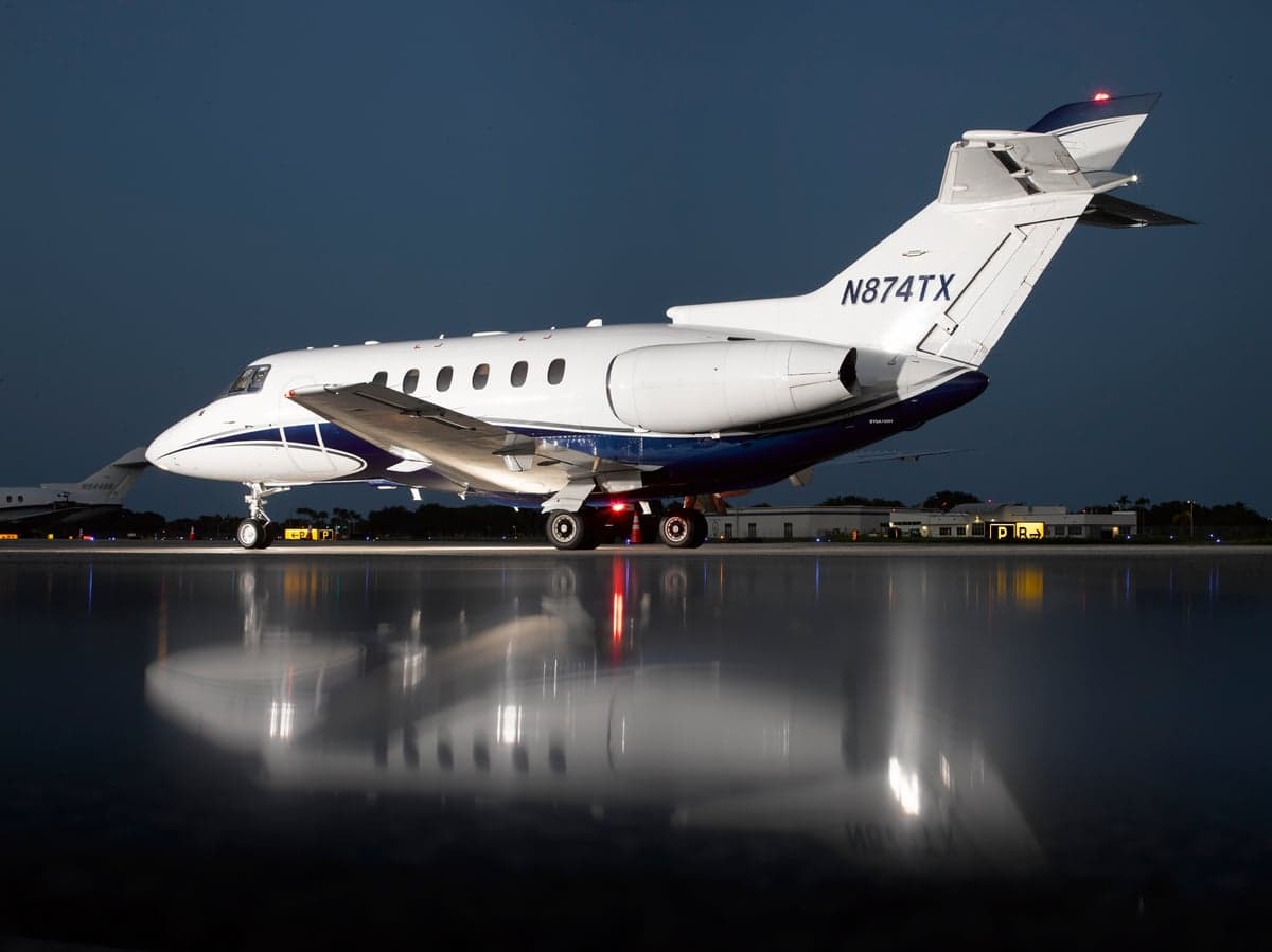 A white and blue business jet (N874TX) is parked on a wet tarmac at night, viewed from the rear.