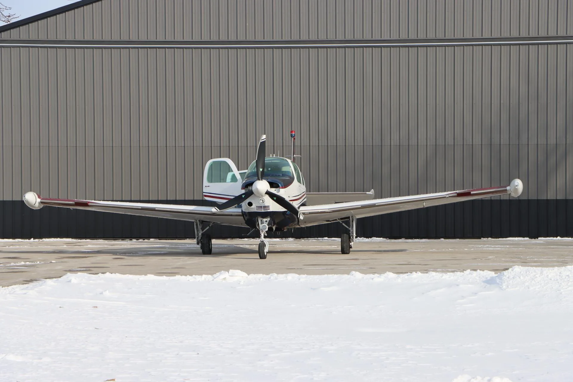 Front view of a white, blue, and red single-engine aircraft with an open door, parked on a snowy tarmac in front of a hangar.