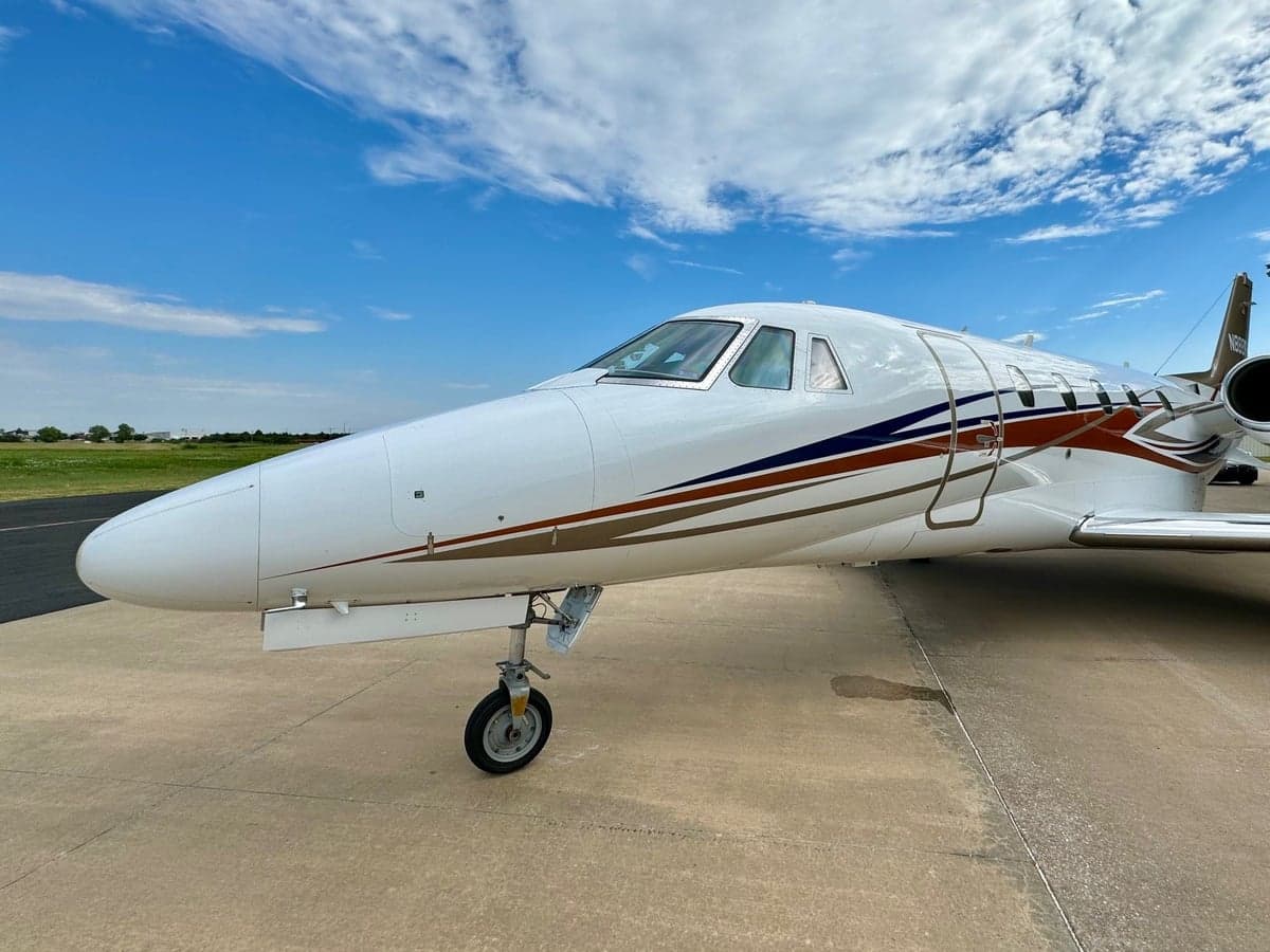 A white business jet with brown, blue, and gold stripes is parked on a concrete tarmac under a partly cloudy sky, viewed from the front-right side.