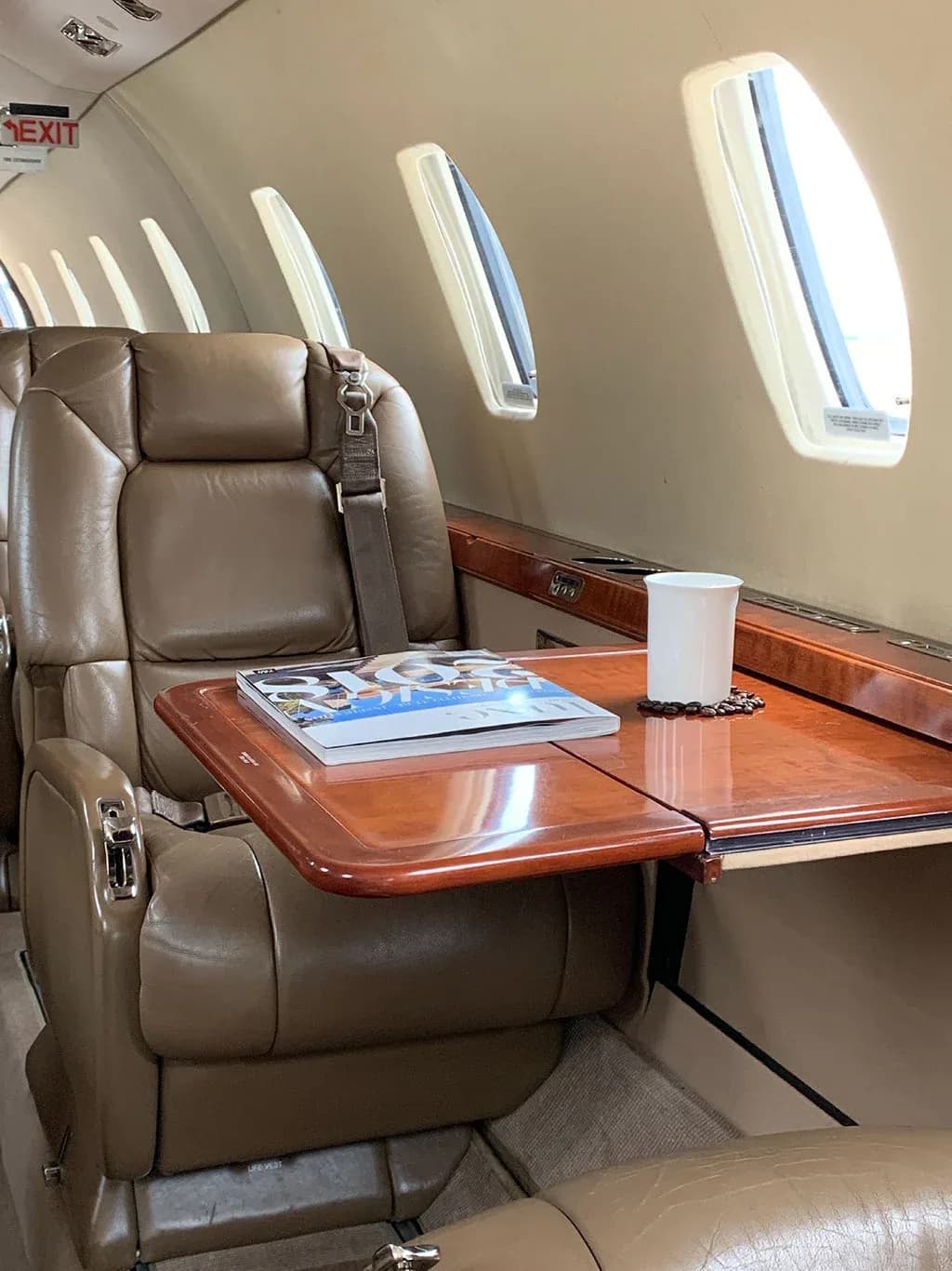 Close-up of a tan leather seat and a fold-out table with a magazine and a white mug in the aircraft cabin.