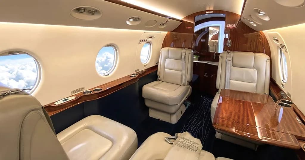 A view of the private jet's cabin with white leather seats, a wooden table, and blue carpet.