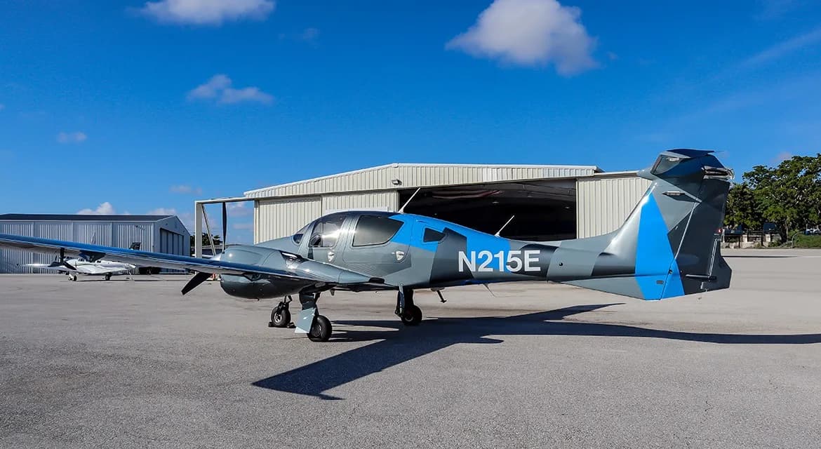 A gray and blue Diamond DA62 aircraft, registration N215E, parked on an airfield tarmac next to a hangar.